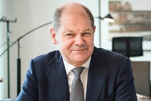 Bundesfinanzminister Olaf Scholz in grauem Anzug, lächelnd, im Hintergrund Arbeitszimmer.