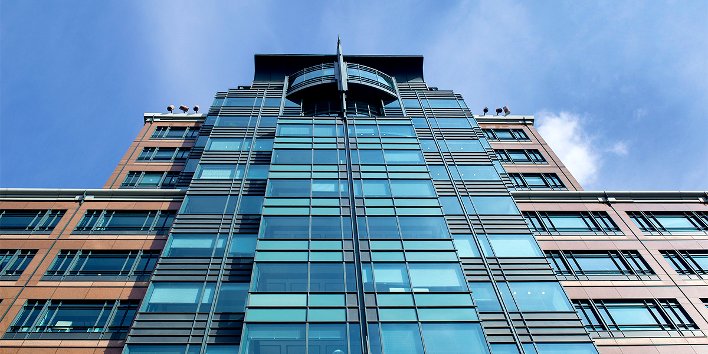 Gläsernes Bürogebäude, von unten gesehen, dahinter blauer Himmel
