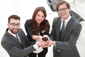 Zwei Jungunternehmer und eine Jungunternehmerin halten zu dritt einen Haufen Erde in den Händen, aus dem eine Pflanze wächst.