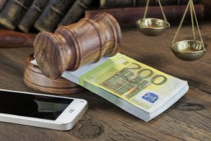 Bündel 200-Euro-Scheine unter Gerichtshammer, davor Smartphone, im Hintergrund Bücher und Waage der Justitia.